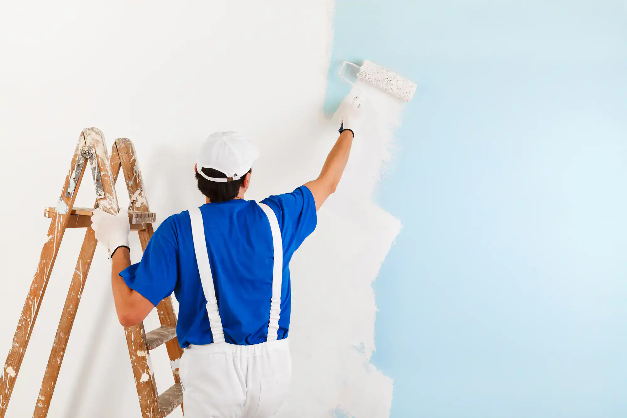 Painter applying white paint to a light blue wall with a roller.