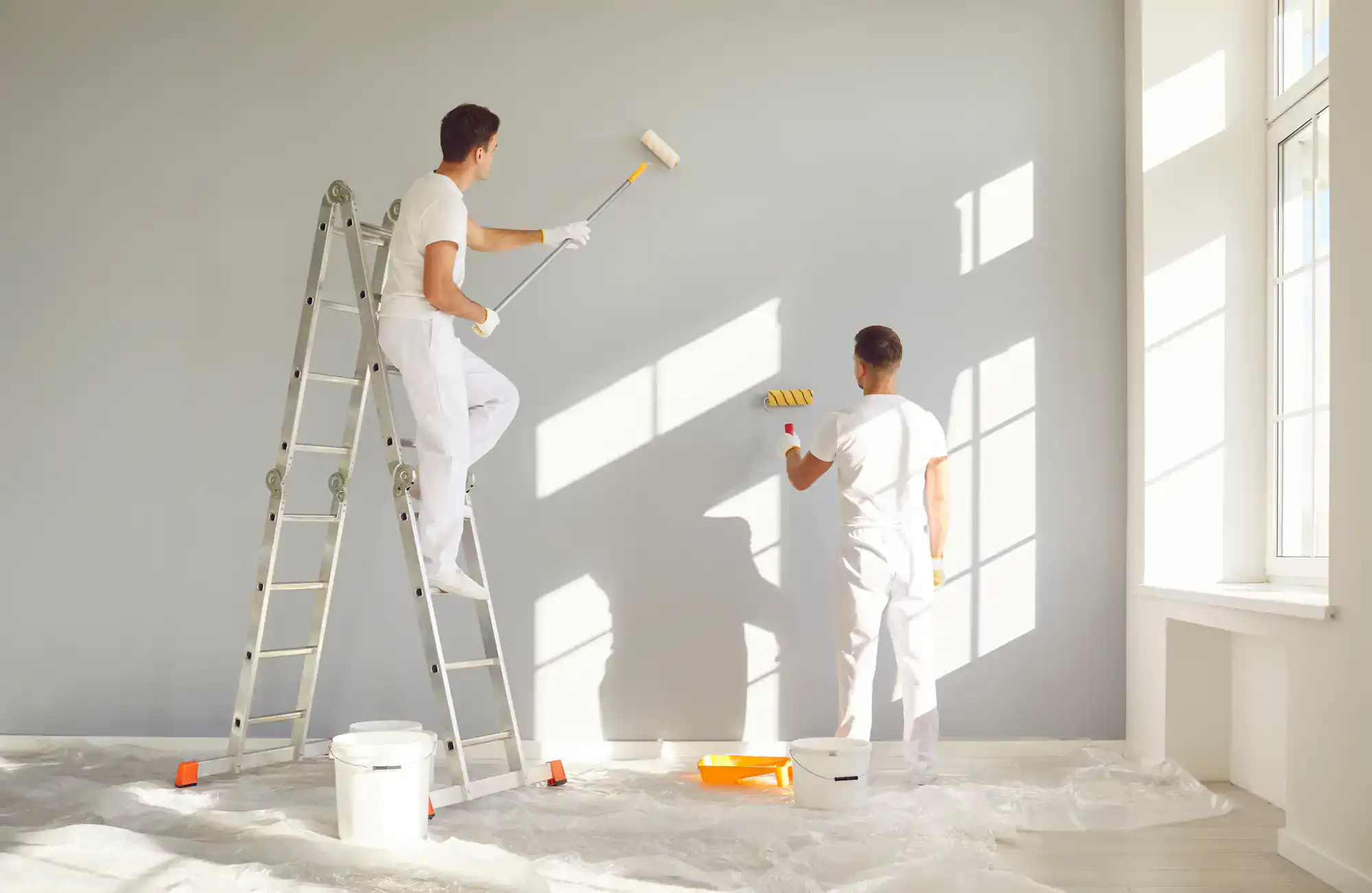 Two painters painting a wall with rollers in a bright room.